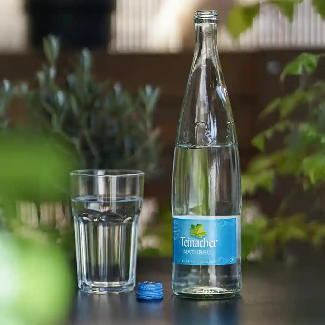 Teinacher Naturell Wasserflasche und Glas auf einem Tisch im Grünen, umgeben von Pflanzen.