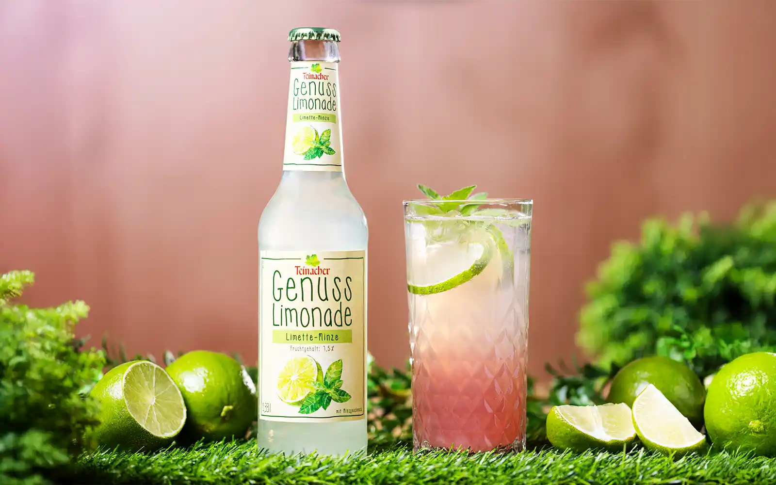 Teinacher Genusslimonade Limette-Minze neben einem Glas Mocktail mit Limettenscheiben und Minze auf grünem Untergrund.