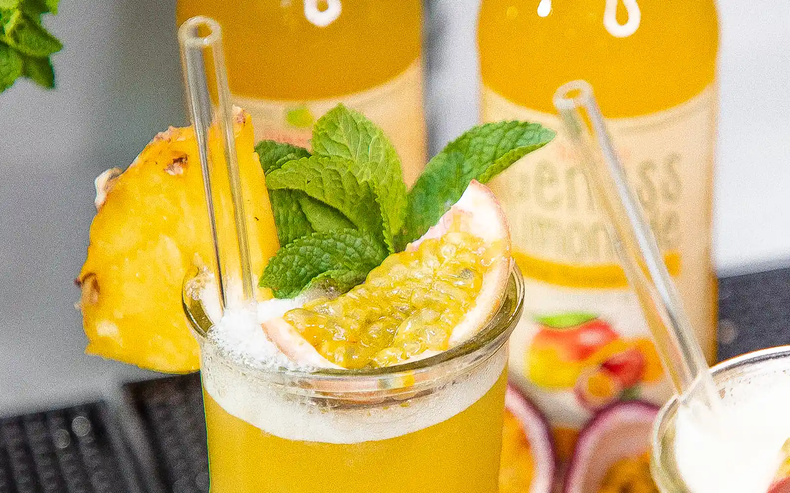 Teinacher Longneck Flaschen Genuss-Limonade Mango-Maracuja-Orange mit 2 Gläsern gefüllt mit Eiswürfeln und Früchten in der Nahaufnahme 